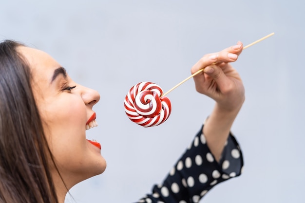 Foto mano della donna che tiene lecca-lecca rossa e bianca. foto ritagliata. donna che prova a mordere la caramella sul bastone