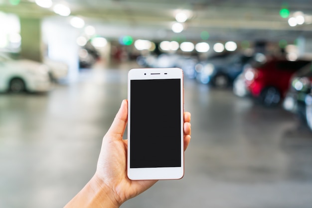 Woman hand holding smart phone with blank screen in blur department store car parking, close up, copy space