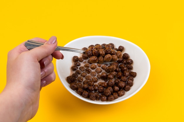 Woman hand holding spoon at White bowl with chocolate corn cereal balls and milk on yellow