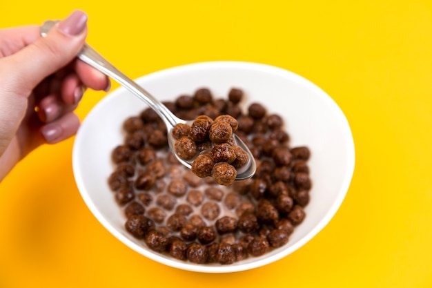 Woman hand holding spoon at white bowl with chocolate corn cereal balls and milk on yellow