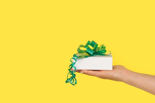 Woman hand holding a white gift box with a green ribbon on a yellow background with copy space