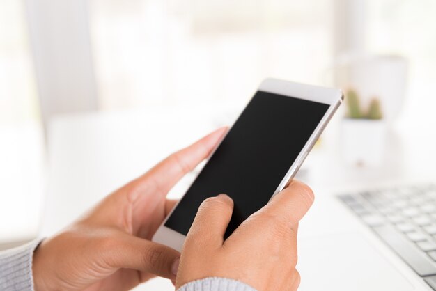 女性の手はオフィスでラップトップを持つテーブルに白い携帯電話を持っています。