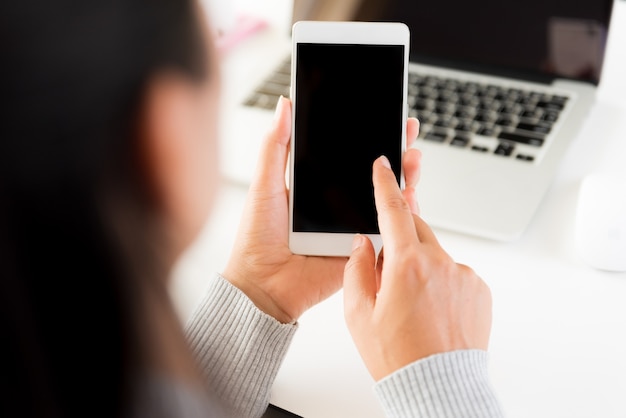 Woman hand holding white mobile phone on a table with a laptop in office