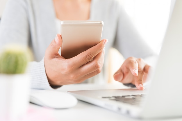 Woman hand holding white mobile phone on a table with a laptop in office