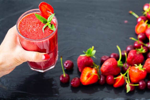 Woman hand holds berries smoothie with strawberries and cherries on black table Selective focus