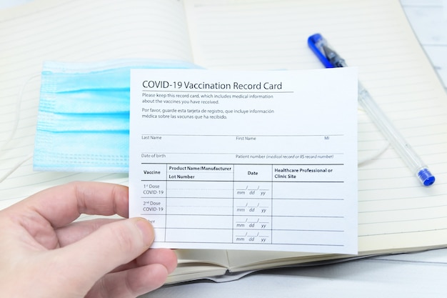 Woman hand holds coronavirus vaccination record card over blue medical mask.