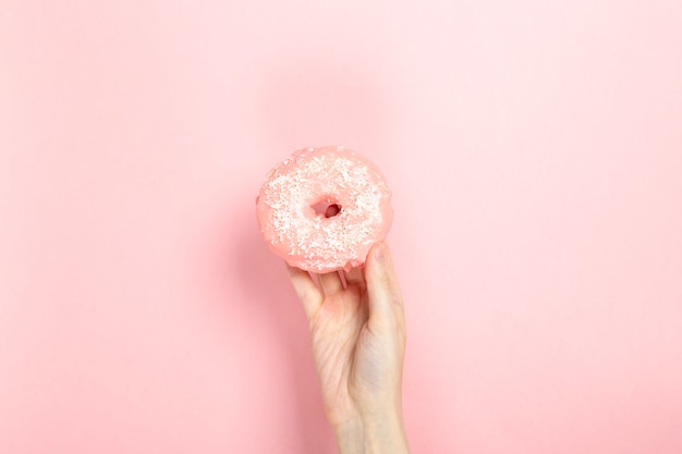 Woman hand holds donut with pink icing and coconut pastry topping, pink background