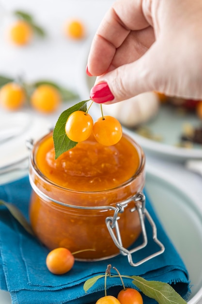 Woman hand holds yellow cherry plum over glass jar of homemade DIY natural canned yellow cherry plum sauce chutney with chilli or tkemali standing on white table selective focus