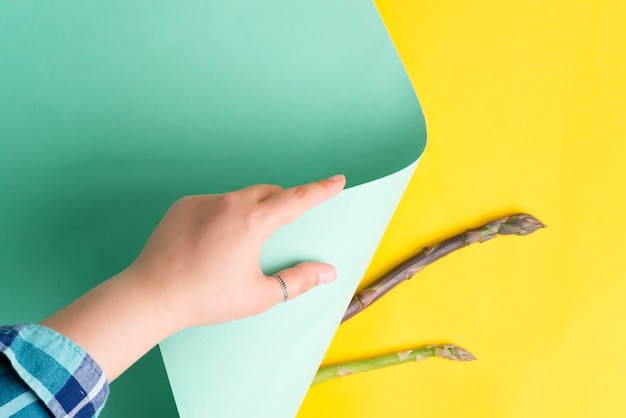 Woman hand is turning pastel turquoise colored paper sheet on a yellow background with new buds of natural asparagus.