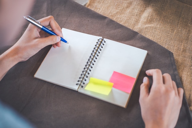 Woman hand is writing on a blank notepad with a pen, on a brown tablecloth.