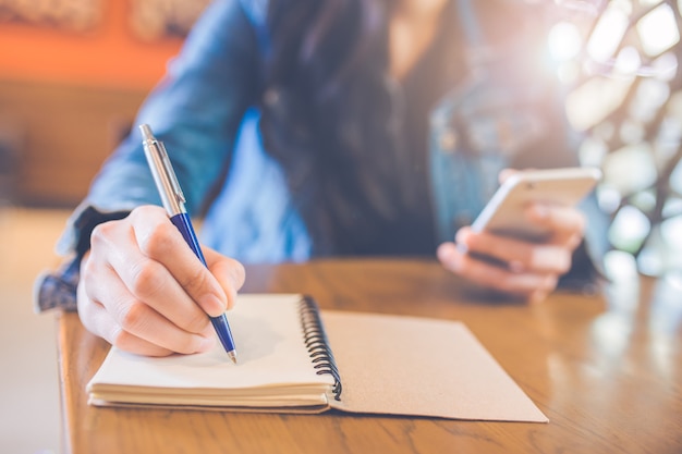 Woman hand is writing in empty spiral notepad with a pen and uses a smartphone.