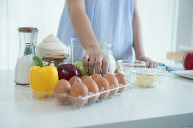 a woman hand in the kitchen in a blue apron takes the egg out of the box.There are apples.