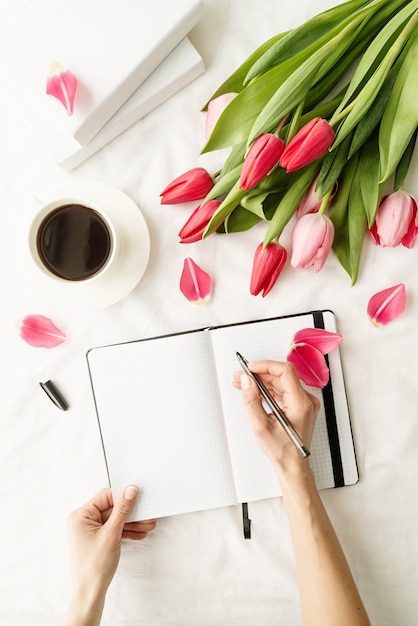 Woman hand making notes in opened notebook, decorated with tulips, coffee cup and books, top view flat lay