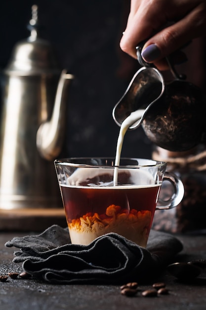 Woman hand pouring milk into glass cup of coffee on black surface