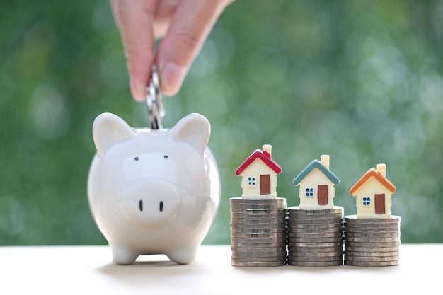 Woman hand putting a coin into piggy bank with model house on coins money on nature green background