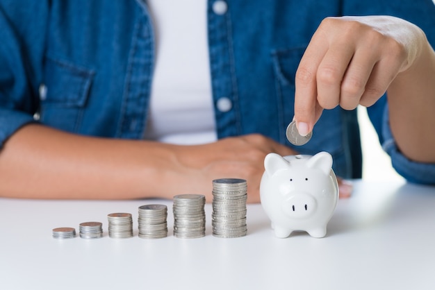 Woman hand putting money coin into piggy bank with stack of coins