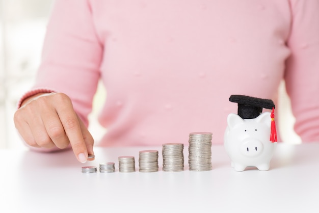 Woman hand putting money coin on stack of coins and piggy bank with graduation cap