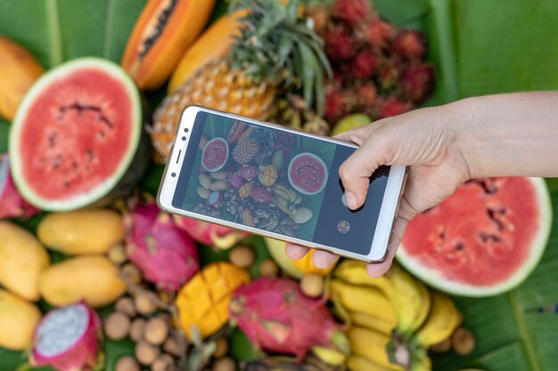 Woman hand take phone photography of tropical fruits Sweet mango papaya pitahaya banana watermelon pineapple Raw vegan vegetarian healthy food