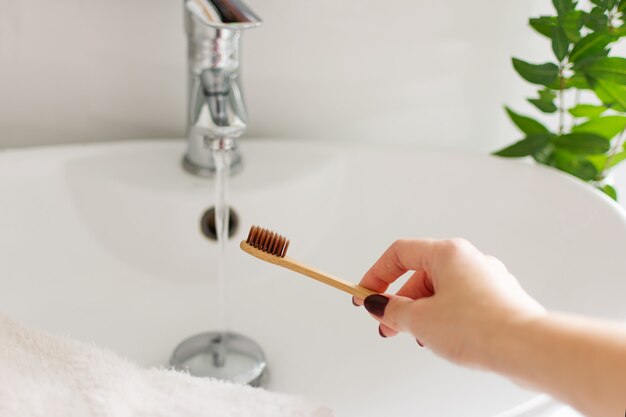 Mano della donna facendo uso del toothbrushe di bambù biodegradabile in un interno bianco del bagno.