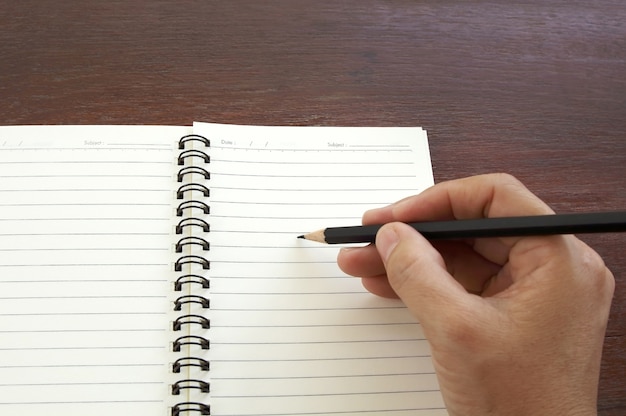 Woman hand with black pencil writing on blank notebook on wooden table.