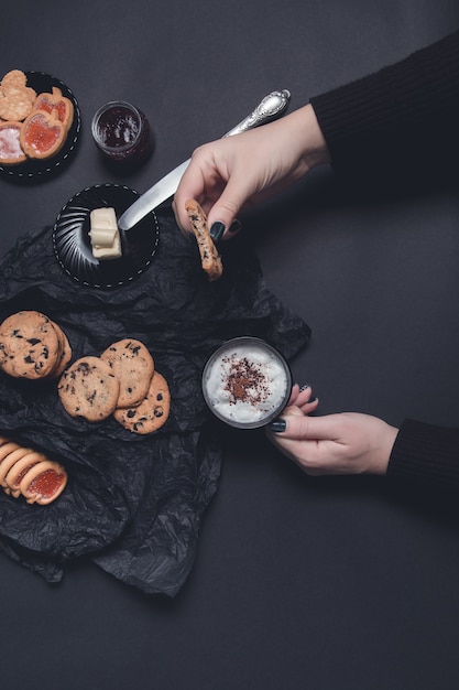 Woman hand with cup of coffee or cappuccino and chocolate cookies, biscuits on black table background. Afternoon break time. Breakfast.