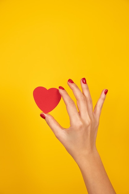 Woman hand with red fingernails holds a paper heart on yellow background Love sign Valentines day concept