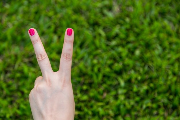Woman hand with red nail show peace sign or number two with two fingers on green grass background