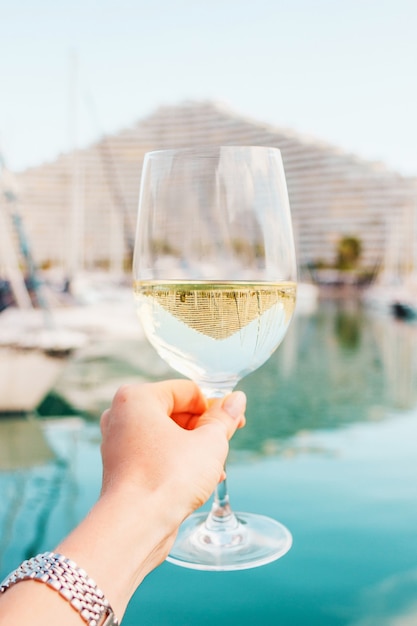 woman hand with white wine champagne glass on yachts sailing boats building background in france