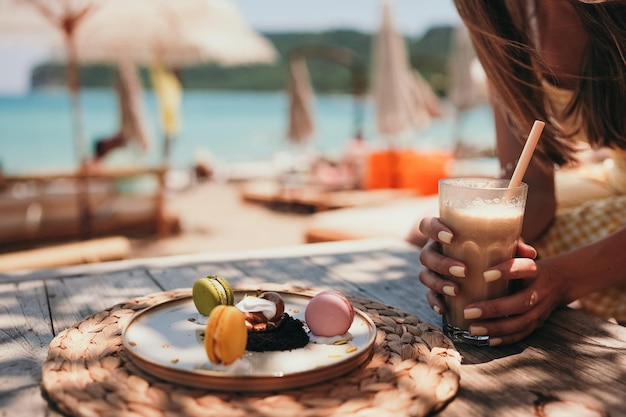 Woman hand with yellow manicure hold ice coffee latte in tall glass with straw by the sea in beach bar