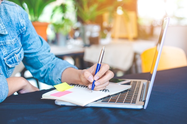 Woman hand works in a laptop computer and is writing on notepad with a pen in the office.