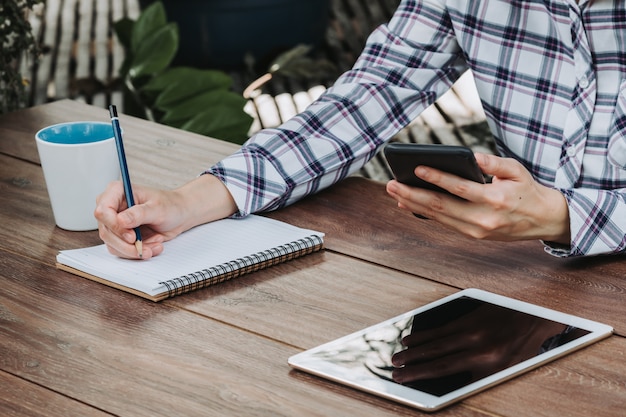 Woman hand writing notepad and holding phone on wooden table at coffee shop