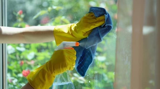 Woman hand in yellow gloves washes window with blue rag and detergent closeup