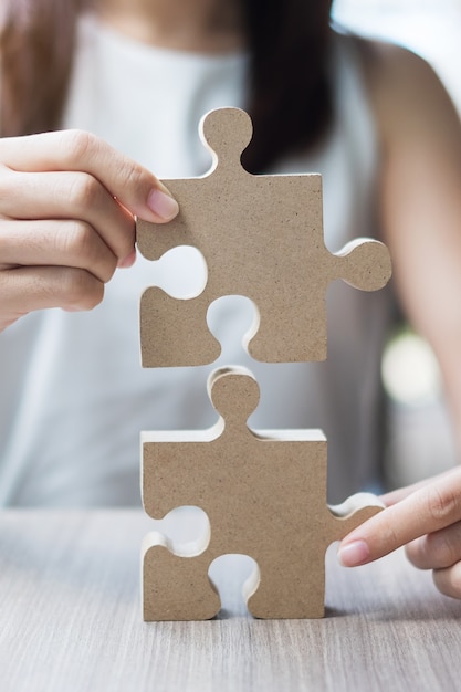 Woman hands connecting couple puzzle over table, businesswoman holding wood jigsaw inside office.