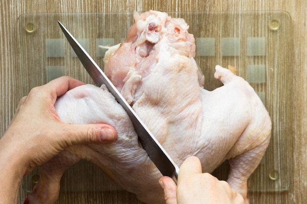 Woman hands cutting off leg with knife from raw body chicken on glass cutting board on wooden background