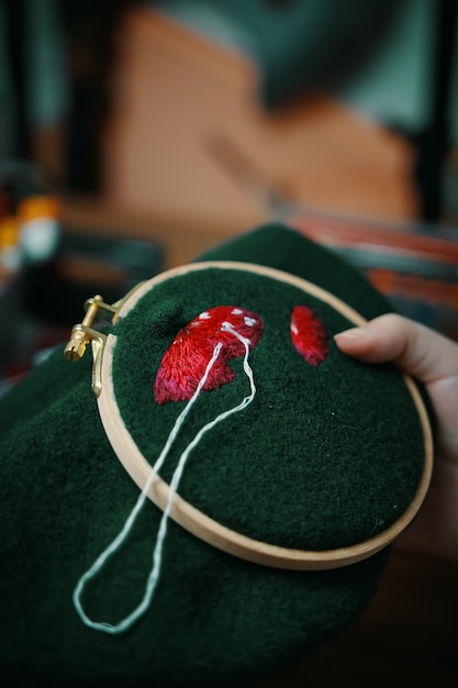 Woman hands doing stitch close up of embroidery of mushroom hat in wooden hoop on green material con...