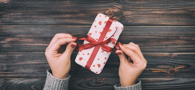 Woman hands giving Holidays gift box on wood table, Present box, red heart decoration of gift on wooden table, top view with copy space for you design. Toned web banner