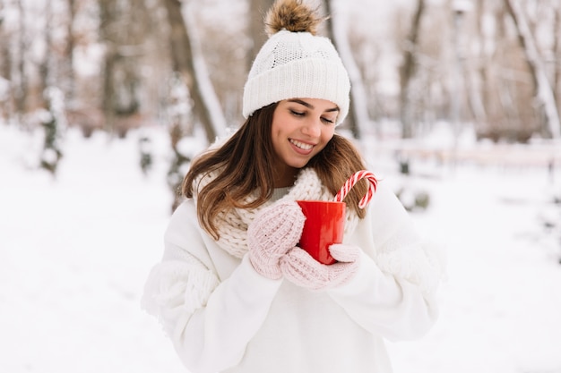 Woman hands in gloves holding a cozy mug with hot cocoa, tea or coffee and a candy cane.