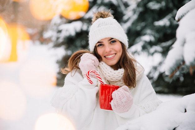 Woman hands in gloves holding a cozy mug with hot cocoa, tea or coffee and a candy cane. 