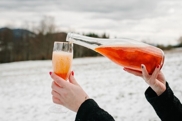 Woman hands hold a bottle champagne and pours into a glass on the background of winter mountains. 