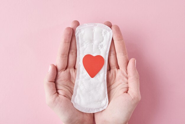 Woman hands hold sanitary pad with red heart on a pink background