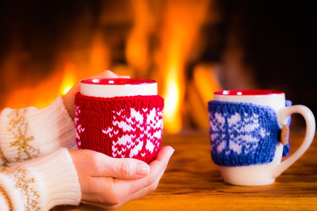 Woman hands holding Christmas cup near fireplace. Winter holiday concept