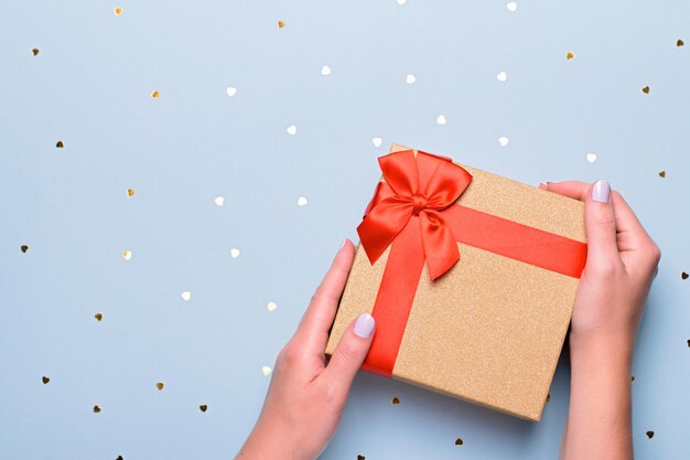Женщина руки держит подарочную коробку в золотых и красных тонах на пастельно-синем фоне с конфетти в форме сердца, copyspace. Открытка на День святого Валентина в модных цветах, С Днем Святого Валентина фон