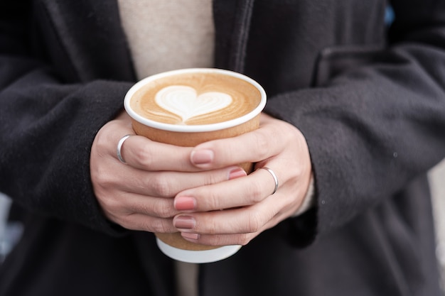 ホットコーヒー紙コップ、ハート形のラテコーヒーアートを保持している女性の手。愛、休日、バレンタインデー、無料のプラスチック容器のコンセプト