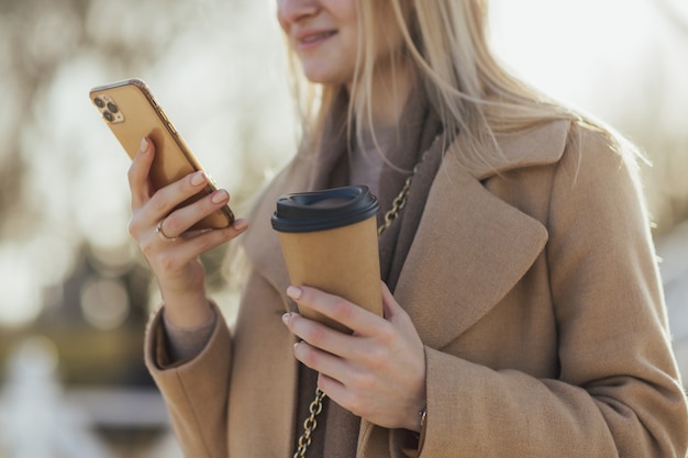 woman hands holding paper cup of coffee and smartphone in the park