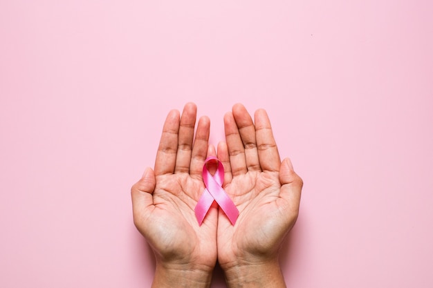 Woman hands holding pink ribbon over pink background, breast cancer awareness, October pink concept