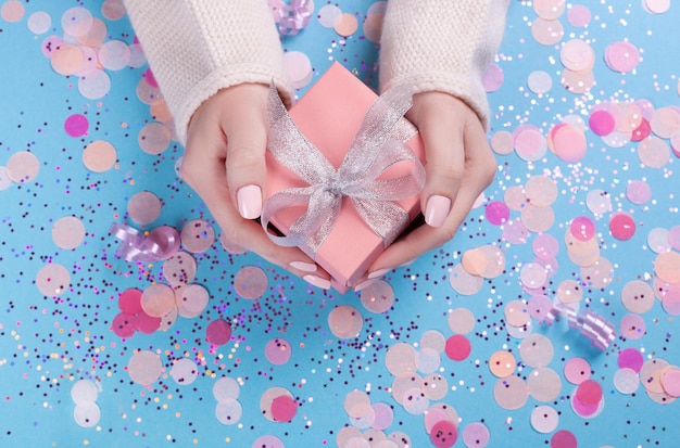 Woman hands holding present box with silver bow on pastel blue background with glitter and confetti