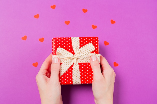 Woman hands holding Red gift present box with small red heart around on purple background. St. Valentine's Day concept. Holiday gift.