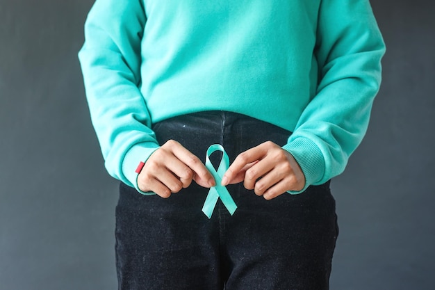 Woman hands holding teal awareness ribbon in front of reproductive organs or uterus Ovarian Cancer