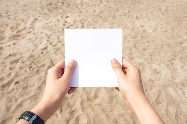 Woman hands holding white paper with sand in the beach texture background.