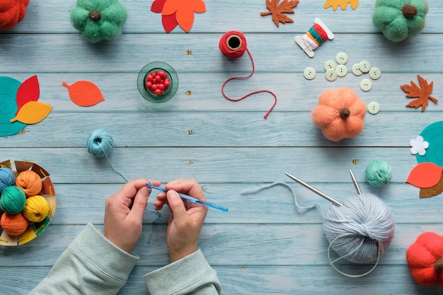 女性の手編みかぎ針編み。糸のボール、ウールの束、装飾的なカボチャ、秋の紅葉と木製のテーブルの平面図です。